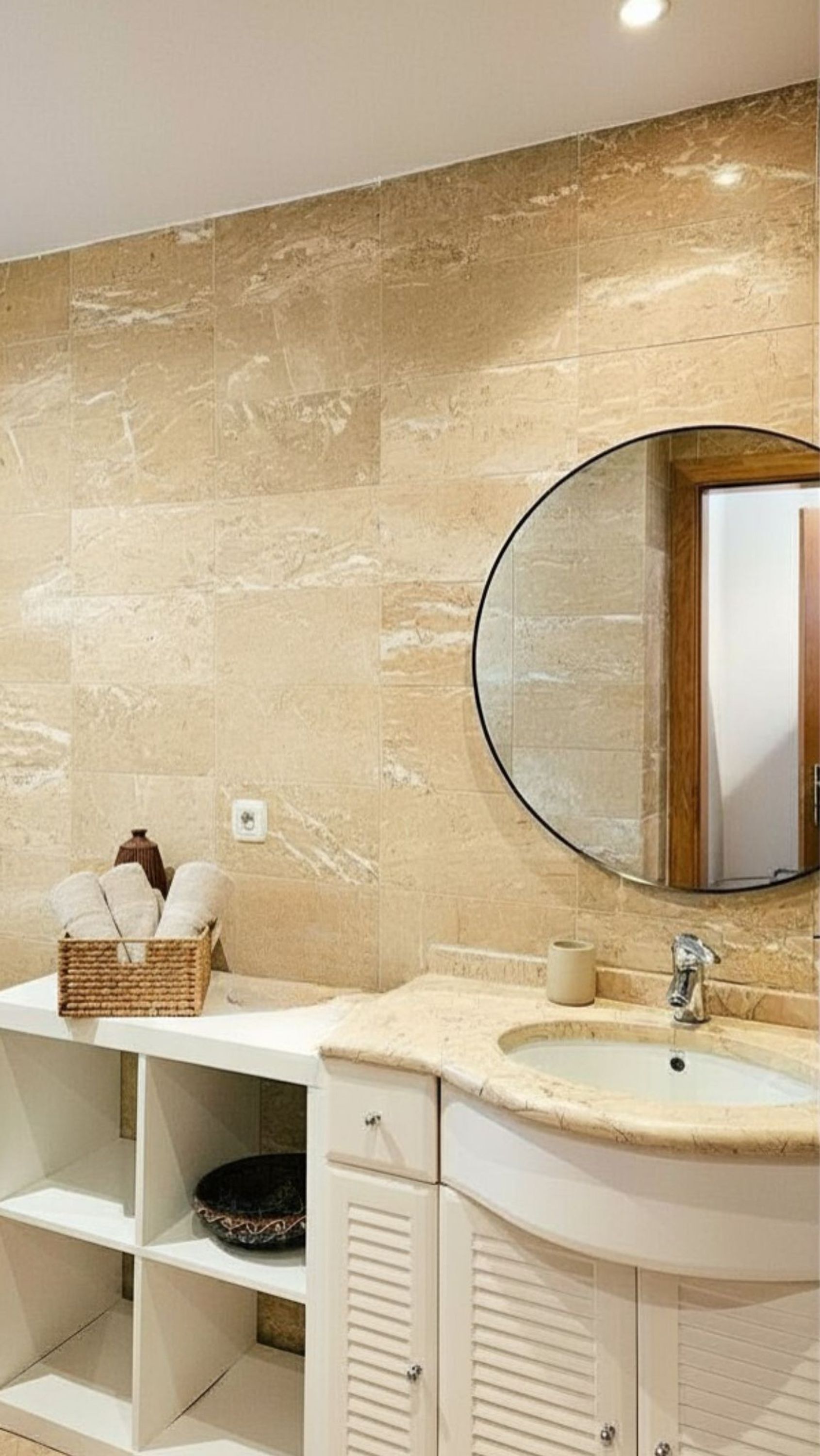 A bathroom with a marble wall, a round mirror, a sink, and a shelf with a basket and a bowl.