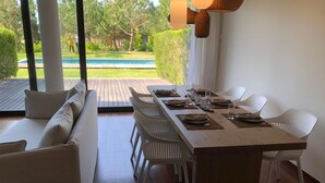 A modern dining area with a long table set for six, white chairs, and a sofa with cushions. - Troia Beach Pool Golf Villa (Grandola)