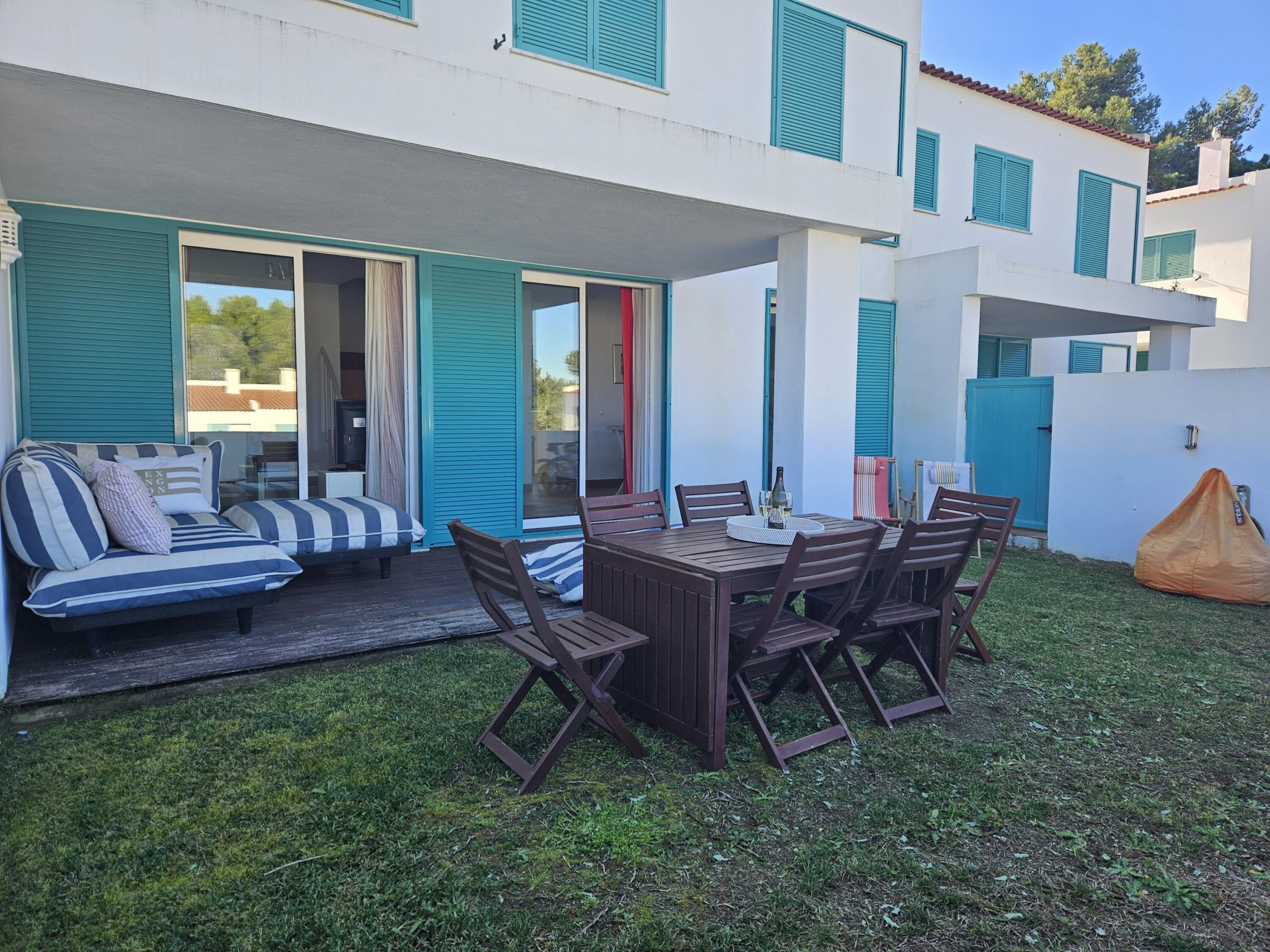 En moderne udendørs terrasse med et træbord og stole, en stribet sofa, og et overdækket siddeområde.