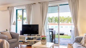 Ein Wohnzimmer mit Flachbildfernseher, einer Couch, einem Couchtisch und Blick ins Freie durch große Schiebeglasüren.