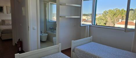 A bedroom with two beds, a window with a view of buildings, a bathroom with a toilet and sink, and a hallway leading to another room.