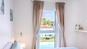 Ein Schlafzimmer mit Bett, einem Fenster mit Vorhängen, einer Nachttischlampe und Blick auf ein Haus und ein Schwimmbad.