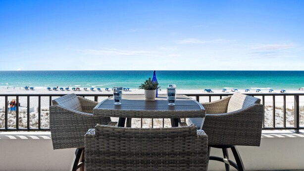 A balcony with wicker chairs and a table, overlooking a beach with umbrellas and a clear blue sky. - SD 410: Seaside Summit - Watch the Sun Sink into the Gorgeous Gulf! (Fort Walton Beach)