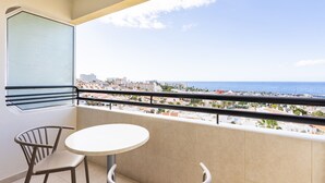Ein Balkon mit rundem Tisch und zwei Stühlen, mit Blick auf eine Küstenstadt mit Gebäuden und dem Meer.