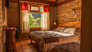 A rustic bedroom with a brick wall, a wooden headboard, a window with red curtains, a bedside table, and a bed with a patterned blanket.