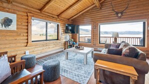 A log cabin interior with a large window, wooden furniture, a mounted animal head, and a view of a lake. - Henry`s Lake View Cabin + BBQ + Fire Pit + Decks! (Island Park)