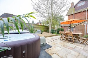 A hot tub in a backyard with a patio table and chairs, surrounded by a wooden fence and a brick house.