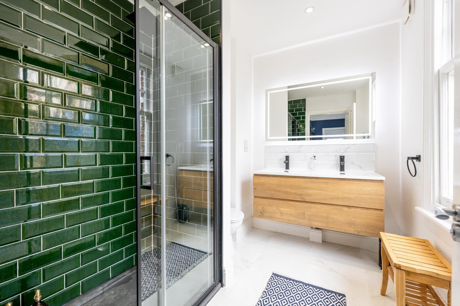 A modern bathroom with a green-tiled wall, a glass shower enclosure, a wooden vanity with a white countertop, and a small wooden stool.