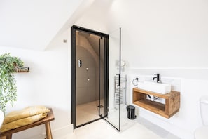 A modern bathroom with a glass shower door, a wooden bench, a sink with a wooden shelf, and a potted plant.