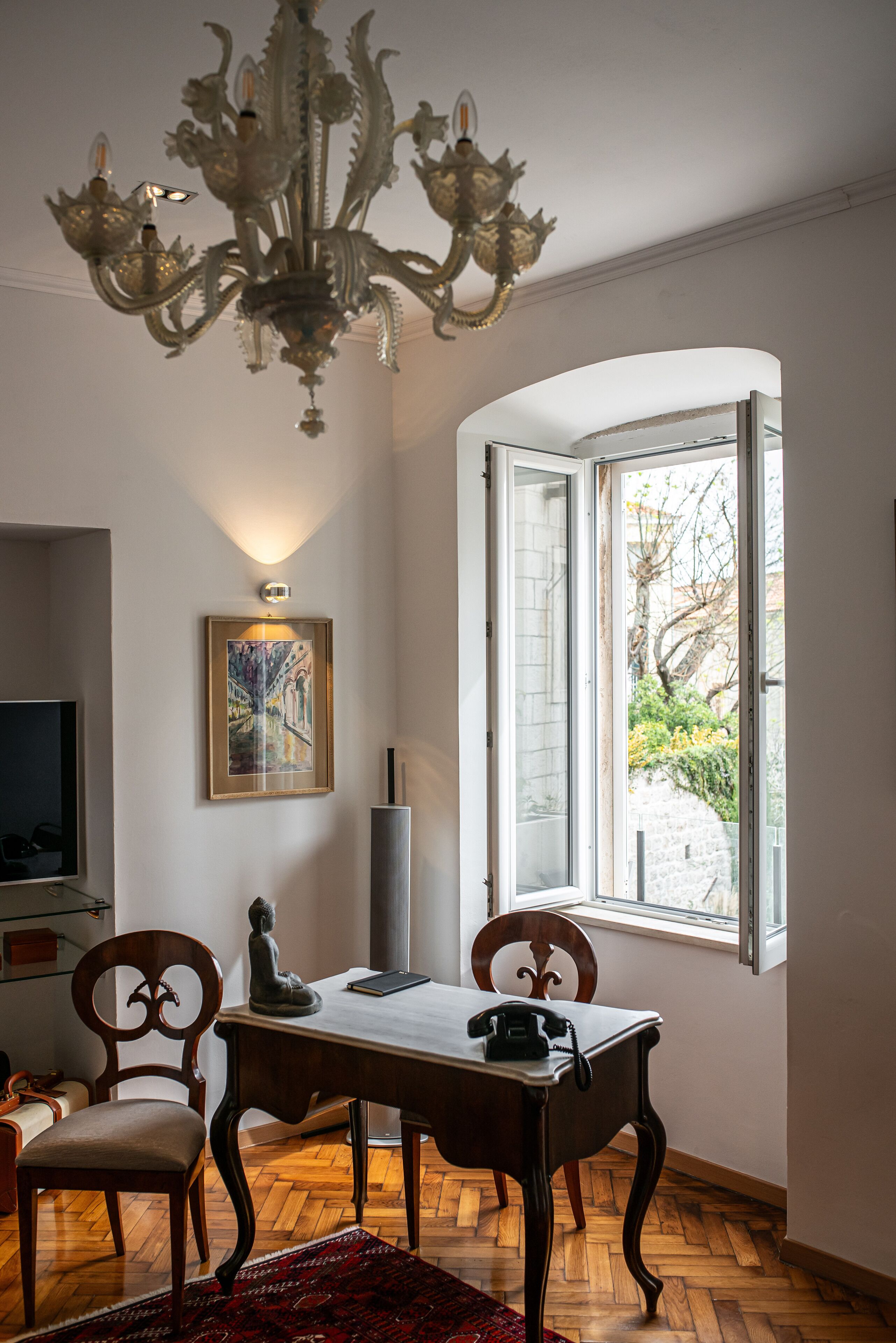 A dining area with a chandelier, a table with chairs, a framed picture on the wall, and a window with a view of the outside.