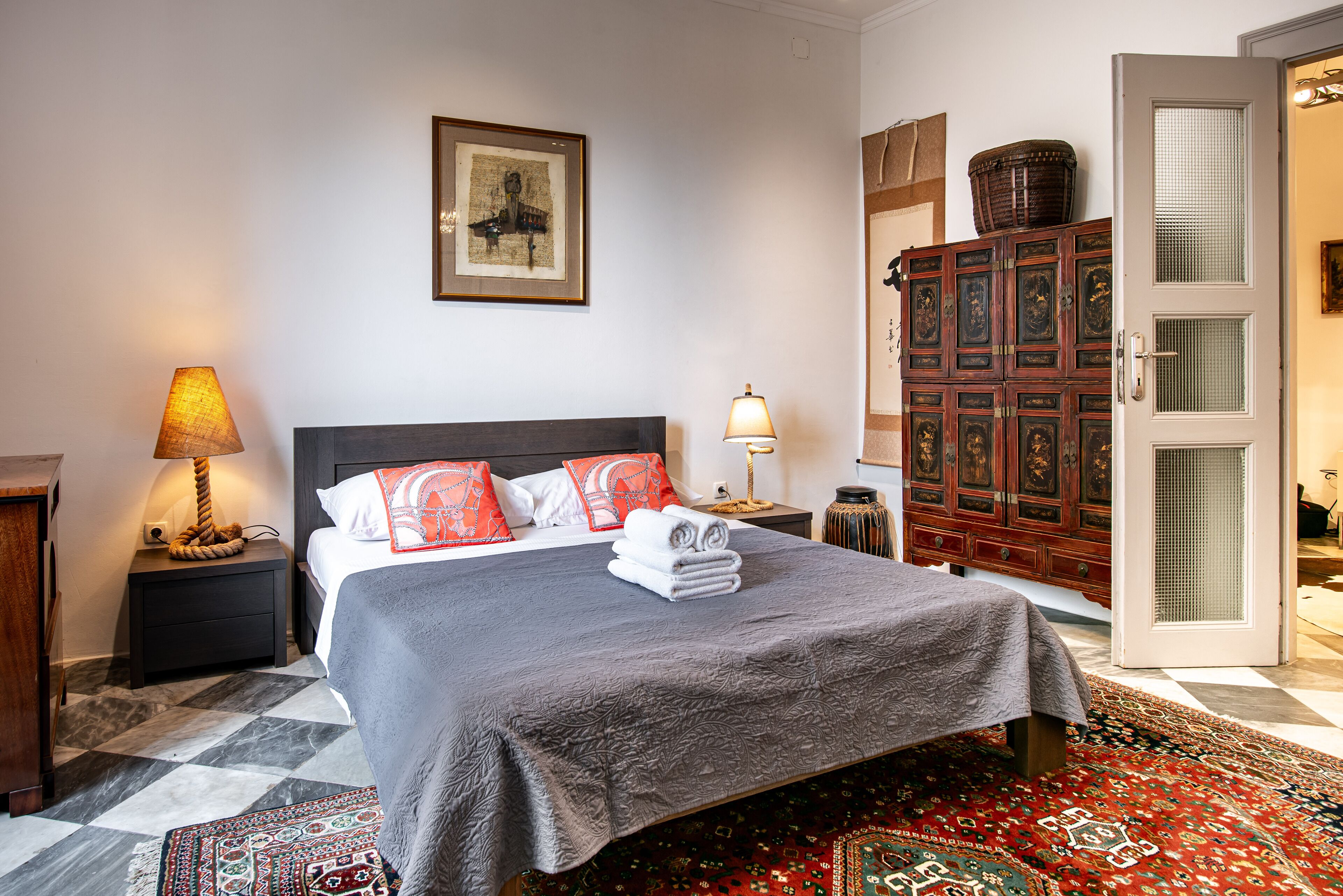 Een net opgemaakt bed met een grijze plaid en rode kussens, een houten nachtkastje met een lamp, een ingelijst foto op de muur, en een klassiek houten kastje.