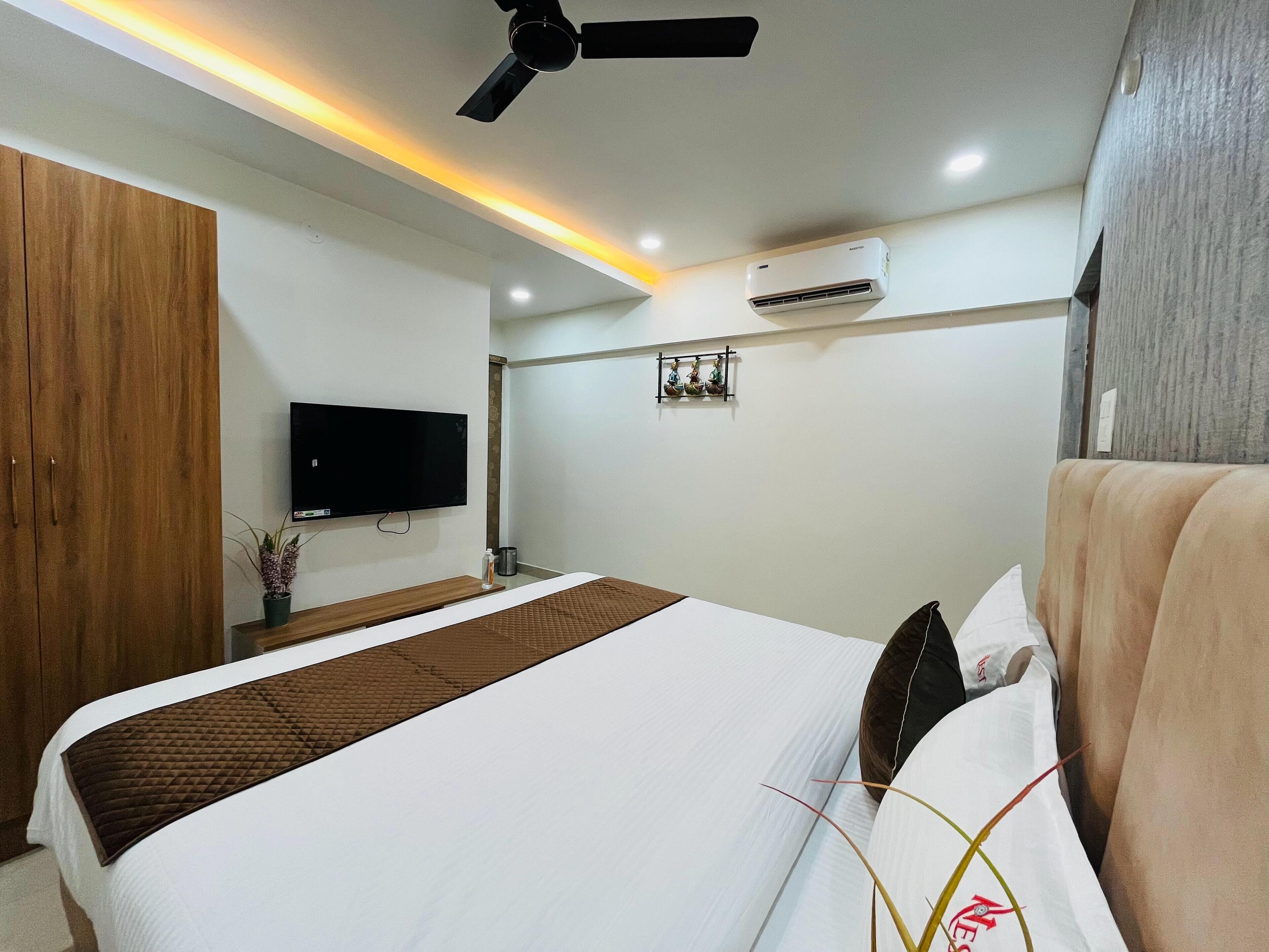 Une chambre d’hôtel équipée d’un lit, d’un ventilateur de plafond, d’un appareil de climatisation, d’un téléviseur à écran plat et d’une petite étagère ornée d’objets décoratifs.