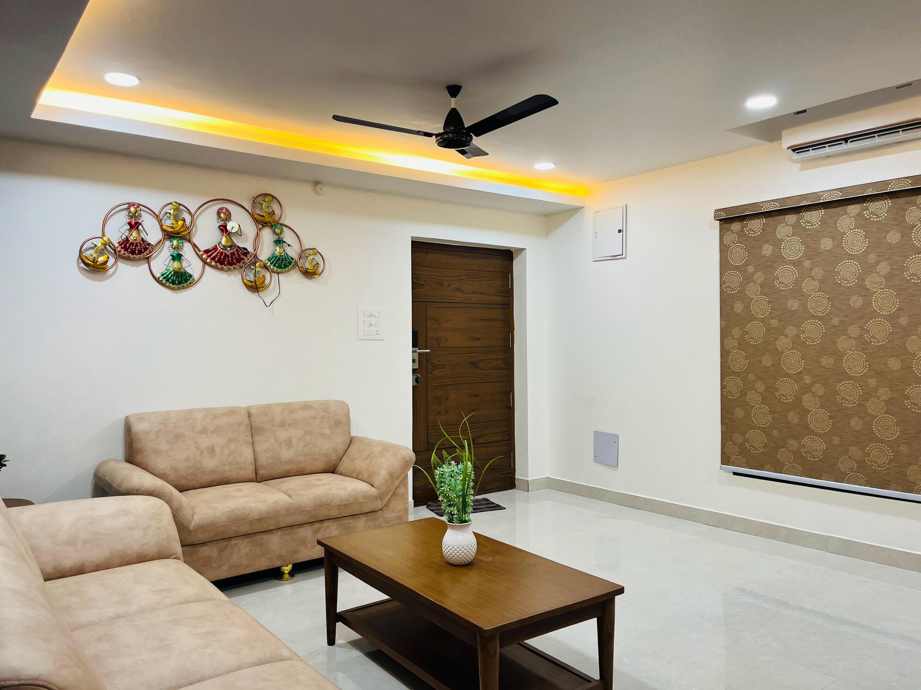 A living room with a sofa, a coffee table with a plant, a ceiling fan, and decorative wall hangings.