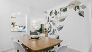 A modern dining area with a wooden table and white chairs, a large leaf-patterned wall mural, and a view of the outdoors through a window. - Luxury Apartment w/ 9 Pools, BBQ & Gym in Cairns (Westcourt)