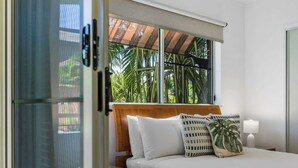 A hotel room with a bed, a nightstand, a lamp, and a window with a view of palm trees. - Luxury Apartment w/ 9 Pools, BBQ & Gym in Cairns (Westcourt)