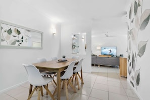 A modern dining area with a wooden table and white chairs, a television in the background, and a grey sofa.