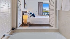 A hotel room with a large bed, a bedside lamp, a view of a balcony, and a bathtub. - Waterfront Apartment with Rooftop Pool and Views (Cairns City)