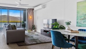 A modern living room with a sofa, a dining table, chairs, a TV, and a view of a lake and mountains. - Waterfront Apartment with Rooftop Pool and Views (Cairns City)