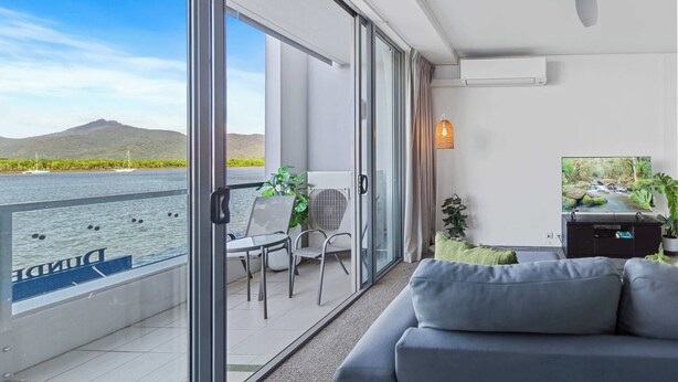 A balcony with a view of a body of water and mountains, a sofa, a TV, and a ceiling fan. - Waterfront Apartment with Rooftop Pool and Views (Cairns City)