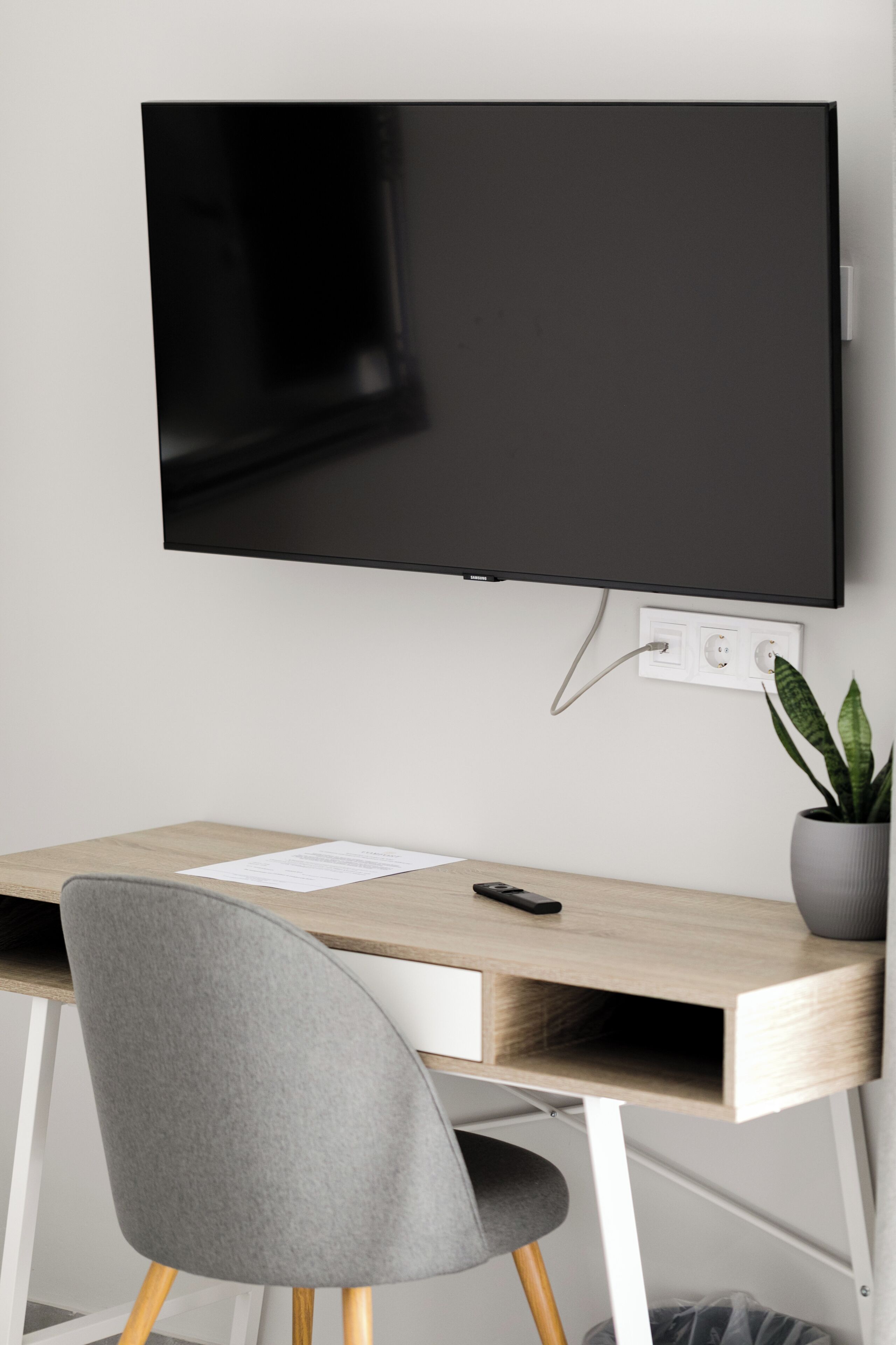 A modern workspace with a flat-screen TV mounted on the wall, a wooden desk, a grey chair, and a potted plant.