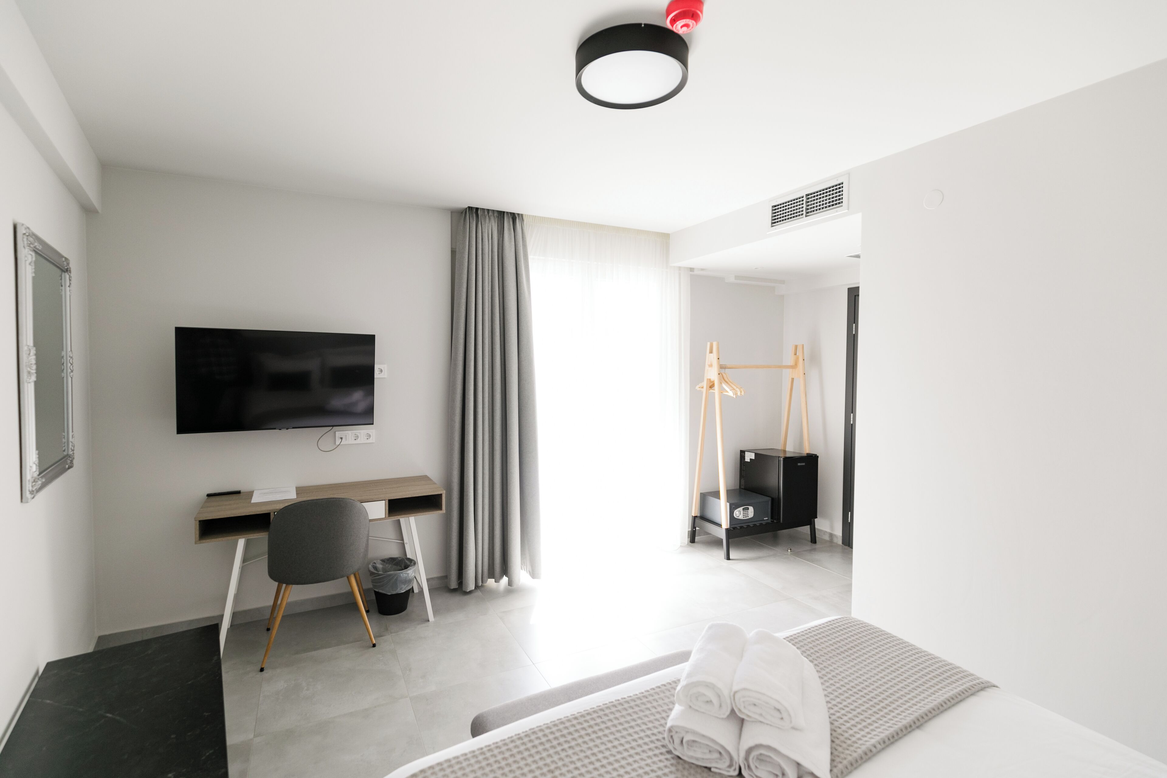 Ett modernt hotellrum med en platt-TV, ett skrivbord, en stol, en säng med hoprullade handdukar och ett stort fönster med gardiner.