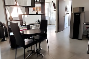 Un comedor moderno con una mesa redonda y sillas negras, una zona de cocina con refrigerador, y vista al exterior a través de una ventana.