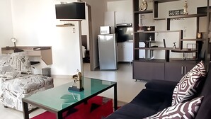 Un salon moderne avec une table basse en verre, un canapé noir et un pouf à motifs. On aperçoit, au fond, une cuisine équipée d’un réfrigérateur et d’un four à micro-ondes.
