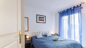 Uma cama bem arrumada com um cobre-leito azul, uma luminária de cabeceira e um quadro na parede.