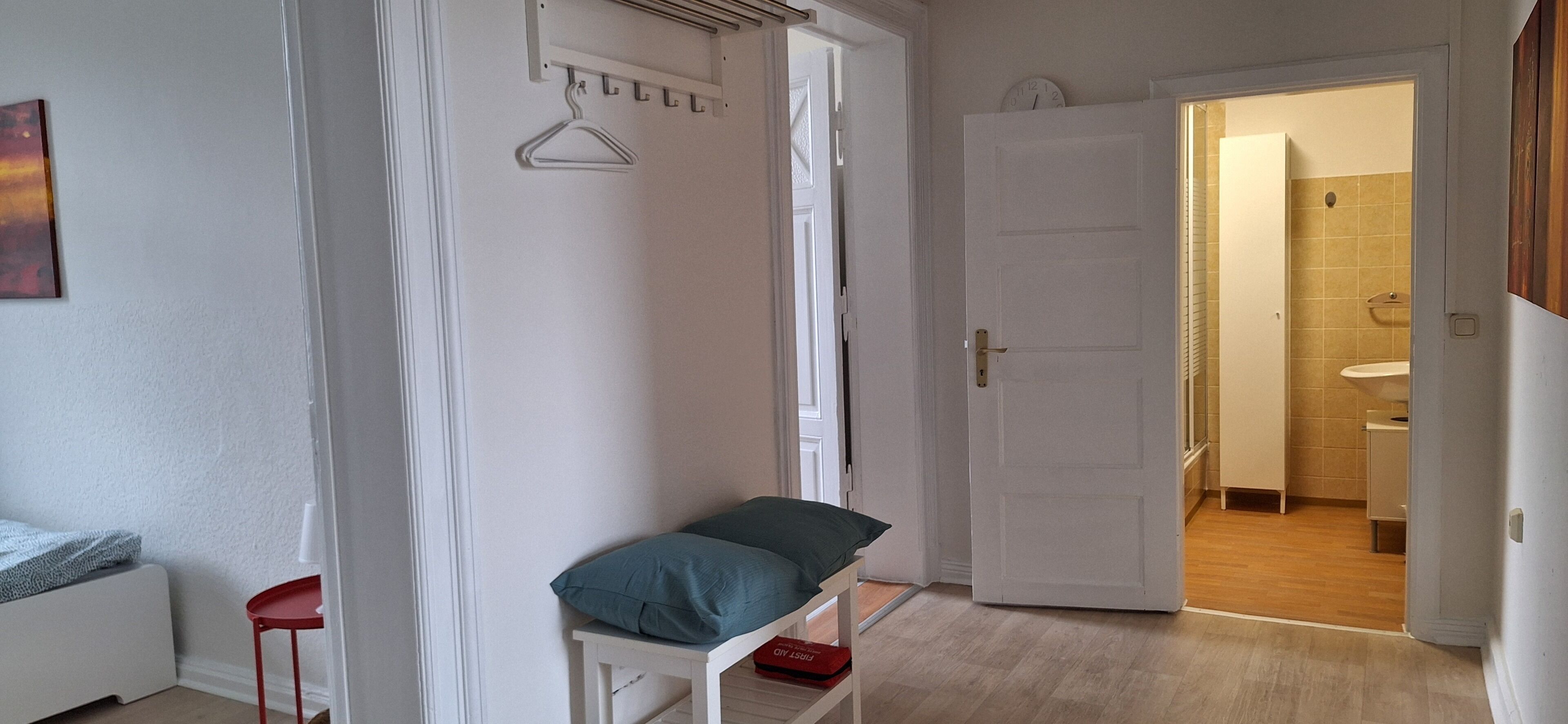 A room with a bed, a small table, a hanging coat rack, and a door leading to a bathroom.