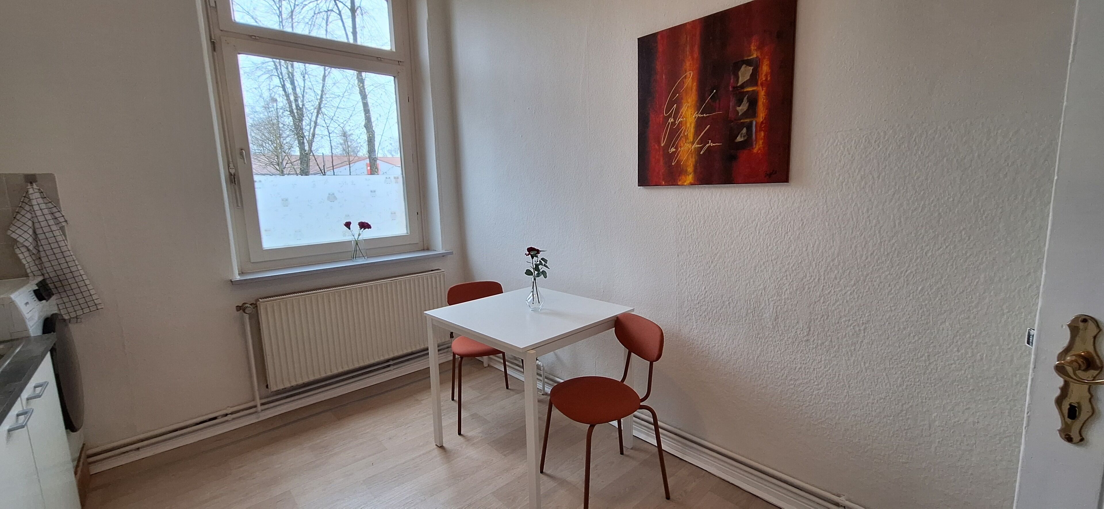 Un comedor con una mesa blanca y sillas rojas, un radiador, una ventana con vistas a árboles y suelo cubierto de nieve, y una obra de arte firmada en la pared.