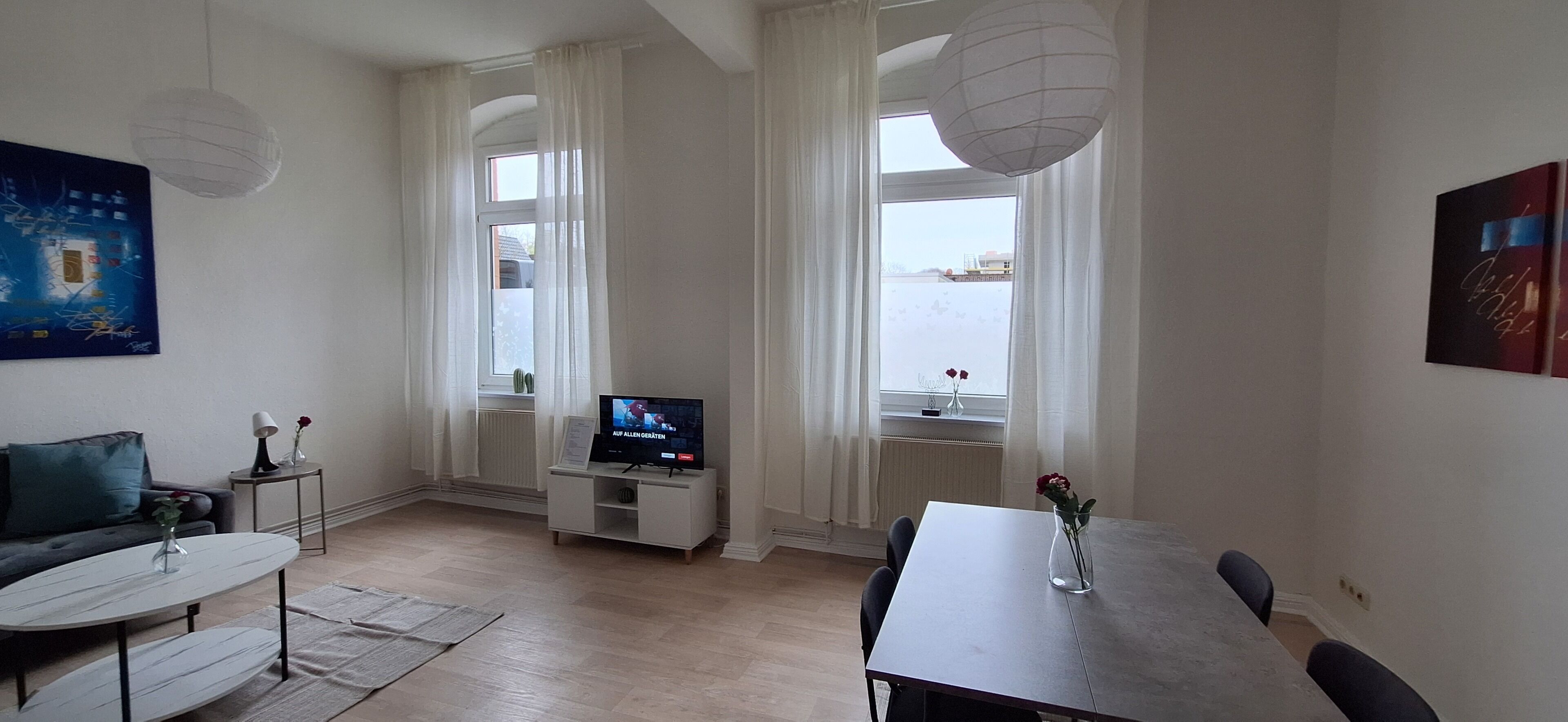 A modern living room with a sofa, a coffee table, a TV on a stand, a dining table with chairs, and large windows with curtains.