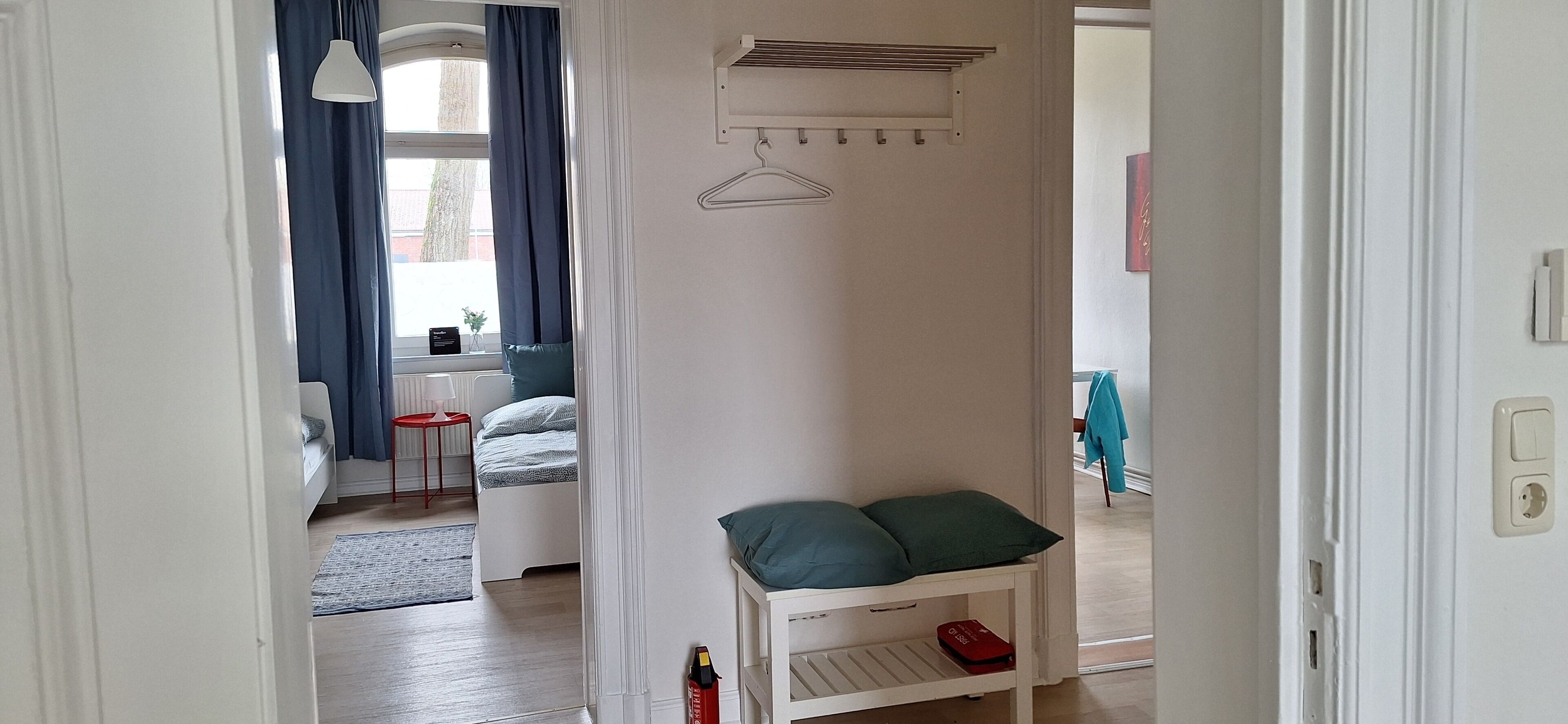 Un couloir étroit mène à une chambre où se trouvent un lit, une petite table et un porte-manteau mural.