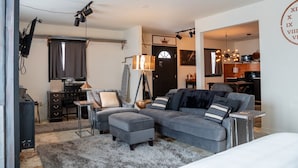 A modern living room with a grey sectional sofa, a small table, and a TV. There is a door leading to a kitchen area. - Orchard Apartment @ The Copper Crown, Bayfield (Bayfield)