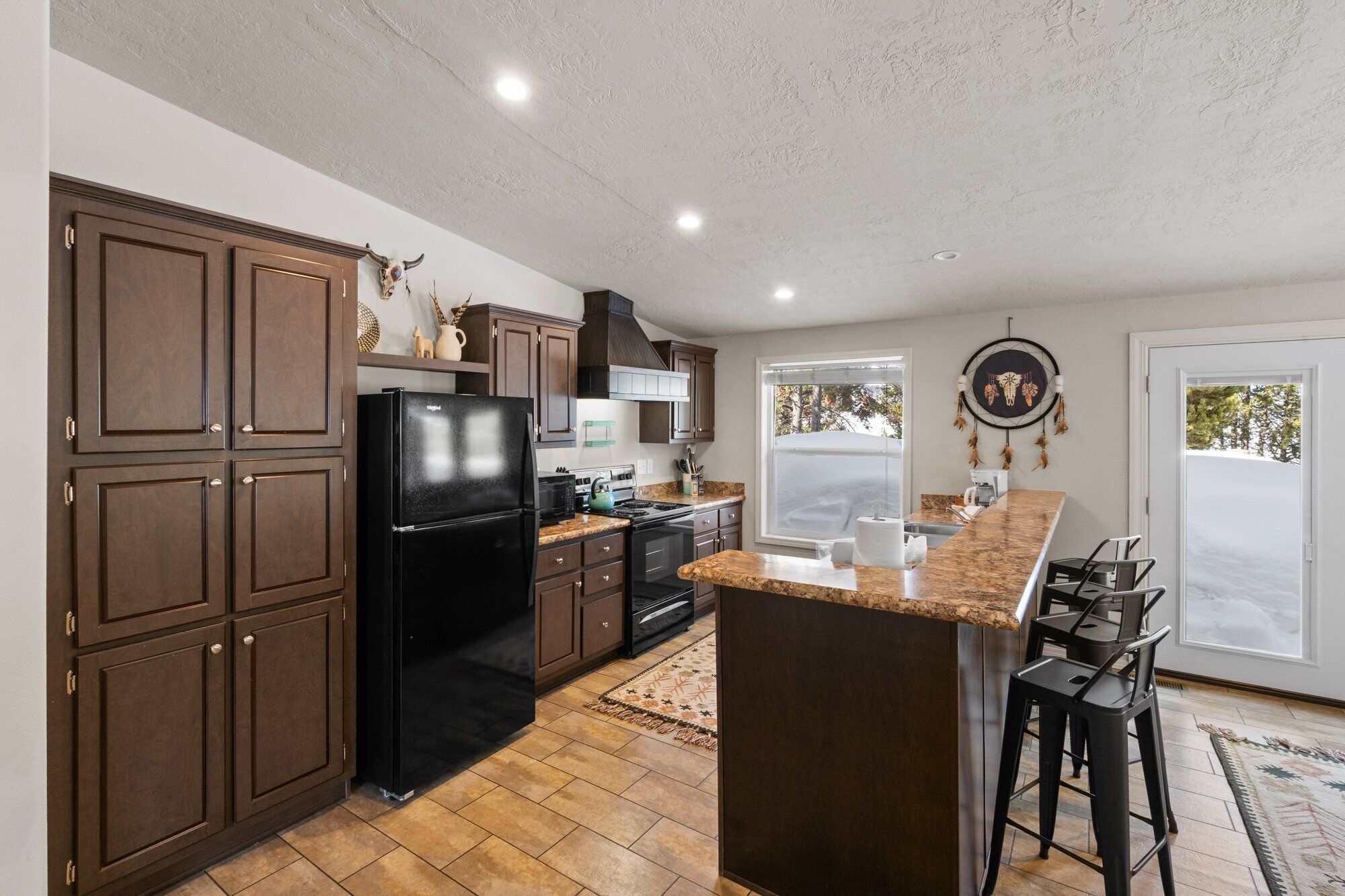 A kitchen with dark wooden cabinets, a black refrigerator, a granite countertop, and a large window.