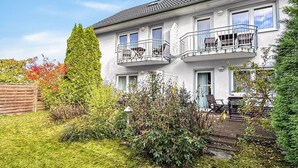 En toetages boligbygning med balkon på hver etage, en hoveddør, samt en lille forhave med et træhegn og forskellige planter.