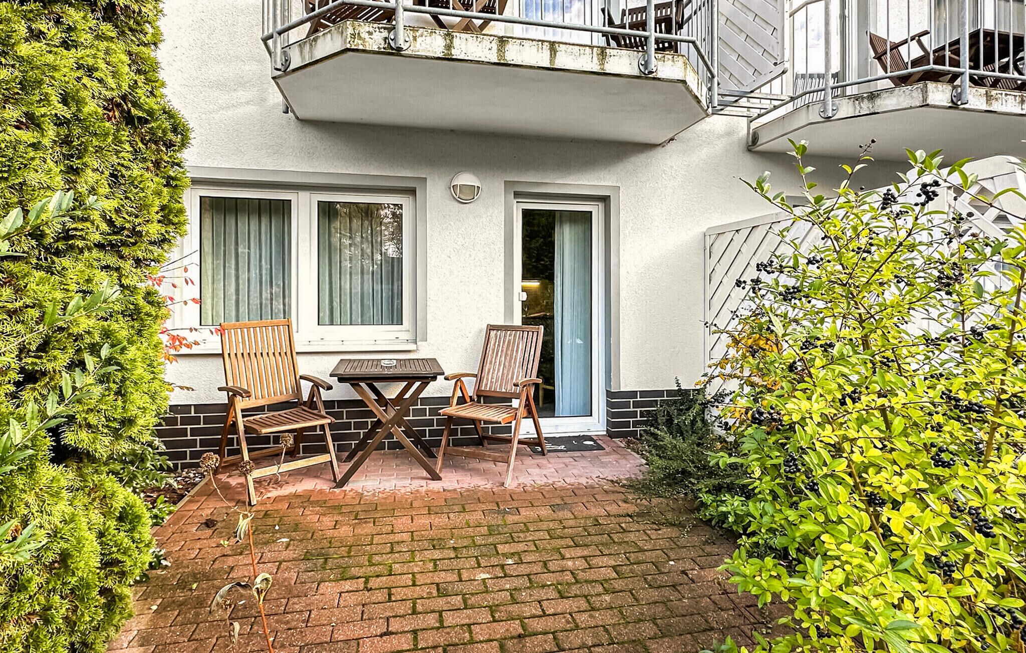 A small patio with two wooden chairs and a table, surrounded by greenery and a brick pathway.