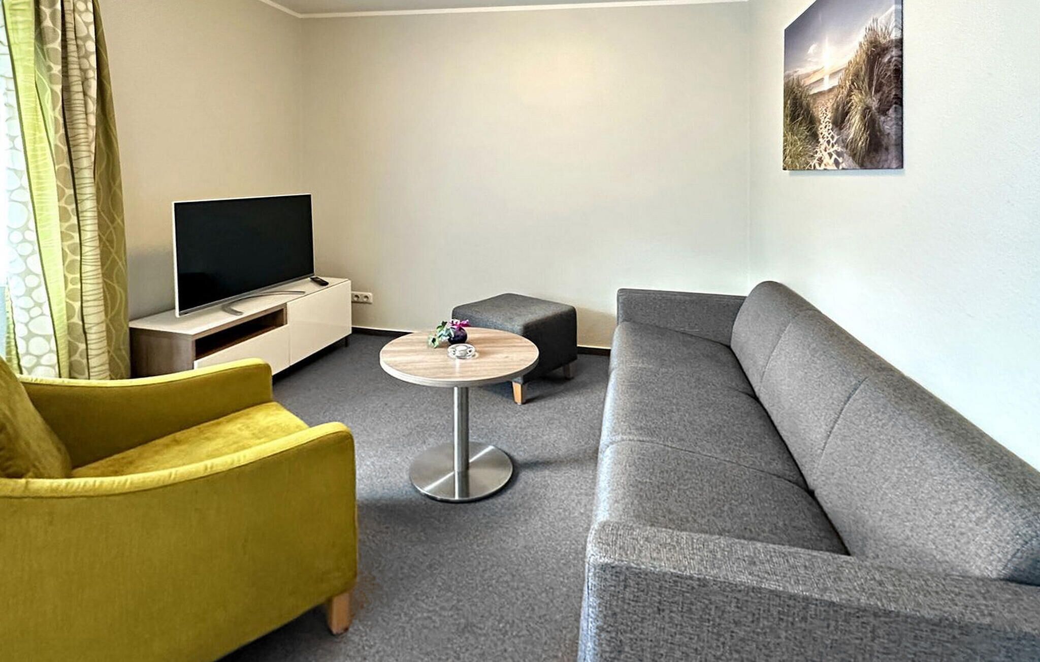 Una sala de estar moderna con un sofá gris, un sillón amarillo, una pequeña mesa redonda y un televisor de pantalla plana sobre un mueble blanco.