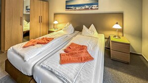 Et dobbeltværelse med to senge, hver med hvide sengetøj og orange håndklæder, to natborde med lamper, et træskab og et maleri af en strandscene på væggen.