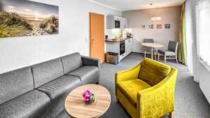 En moderne stue med en grå sofa, en gul lænestol og et lille træbord med en blomst. I baggrunden ses et køkken med et spiseområde og et stort vindue.
