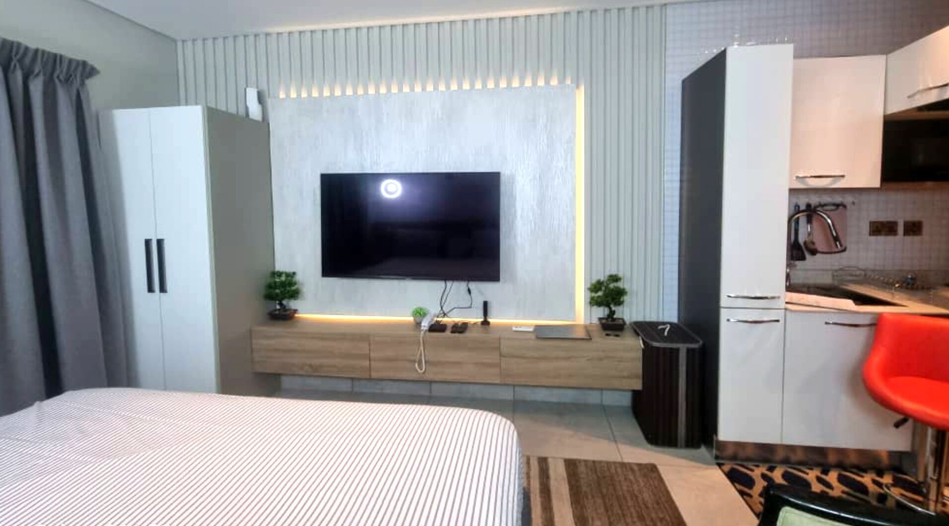 A modern hotel room with a large bed, a TV mounted on a wooden console, a small kitchen area, and a red chair.