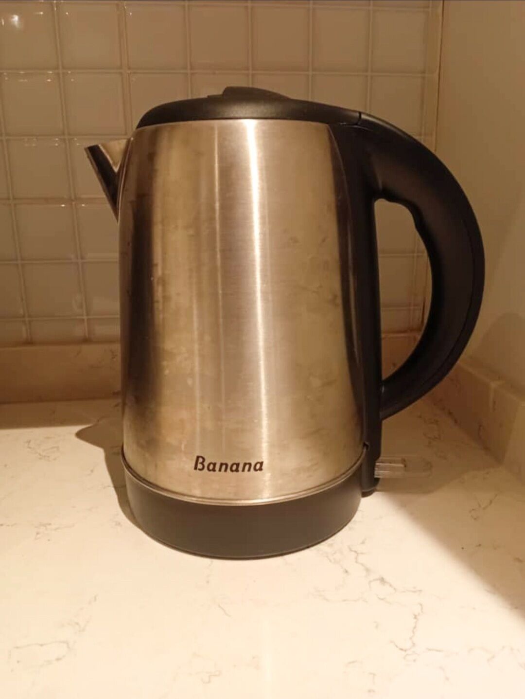 A stainless steel kettle with a black handle and spout, labeled "Banana," on a marble countertop.