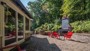 Eine Terrasse im Freien mit roten Stühlen, einem Backsteinboden und einem Kamin aus Stein, umgeben von Grünpflanzen.