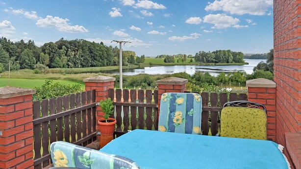 Ein Balkon mit Blick auf einen See und viel Grün, mit einer Backsteinmauer, einem Holzzaun und einem Tisch mit Stühlen.