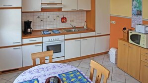 Eine kleine Küche mit weißen Schränken, einem Herd, einer Spüle und einer Mikrowelle. Ein Tisch mit Blumenmuster bedeckt und zwei Stühle stehen bereit.