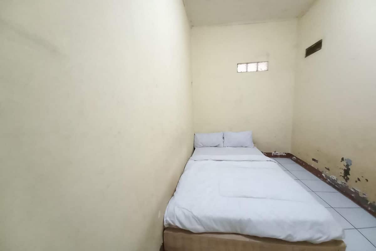 Ein schmales Zimmer mit einem Einzelbett, einer schlichten weißen Bettdecke und einem kleinen Fenster.