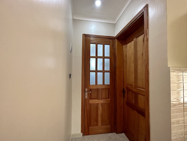 Un pasillo con una puerta de madera y un ventanal de cristal satinado, paredes blancas y suelo de baldosas.