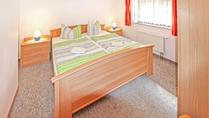 A bedroom with a wooden bed, striped bedding, a nightstand with a lamp, a dresser, and a window with curtains. - Ferienwohnung Mönchgut (Mönchgut OT Alt Reddev)