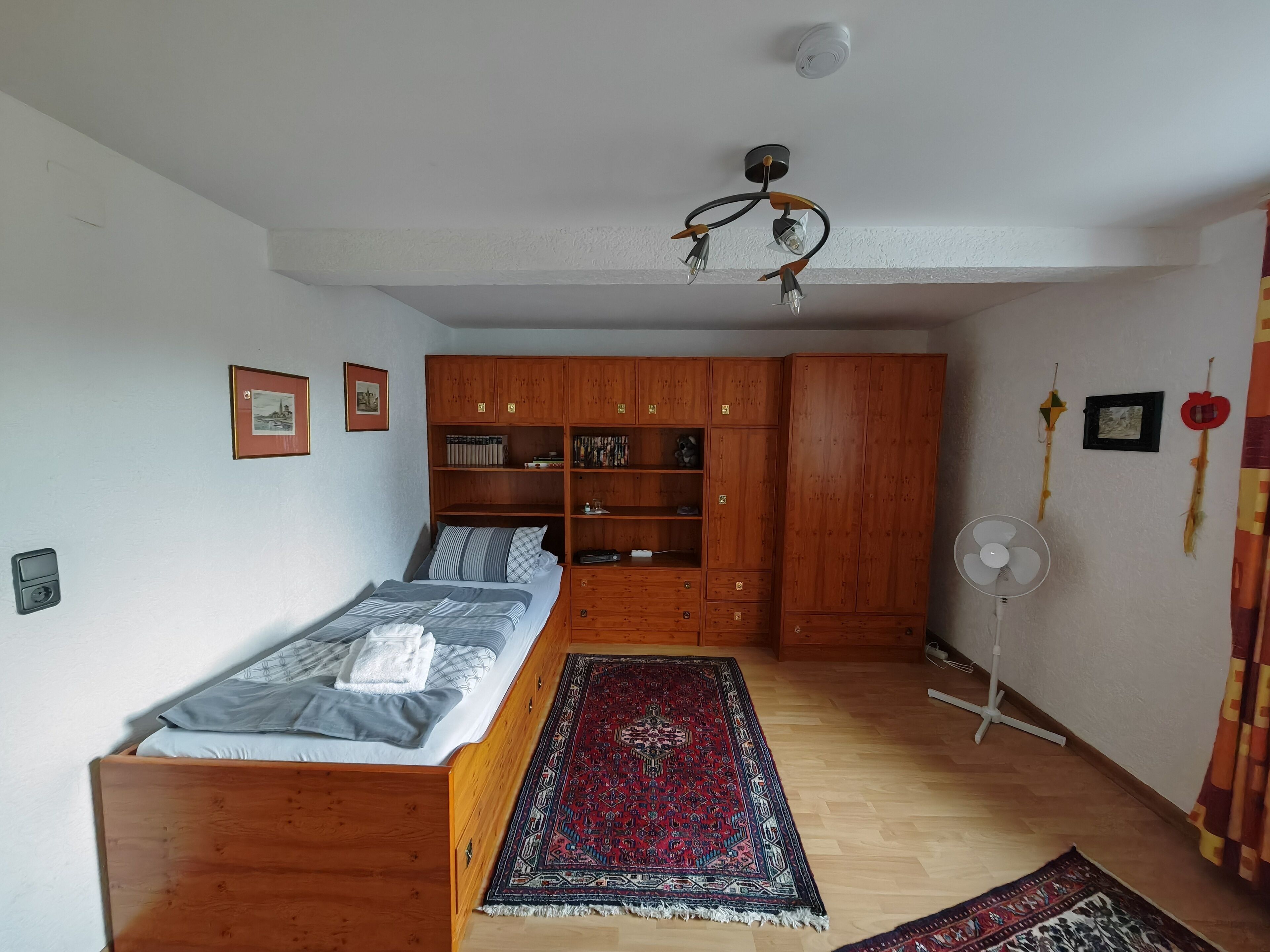 Une chambre à coucher comprenant un lit, une armoire en bois, des étagères, un tapis, un ventilateur et des éléments de décoration murale.