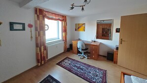 Une pièce comprenant un bureau en bois, une chaise, un radiateur, une fenêtre avec des rideaux, un miroir et une porte.