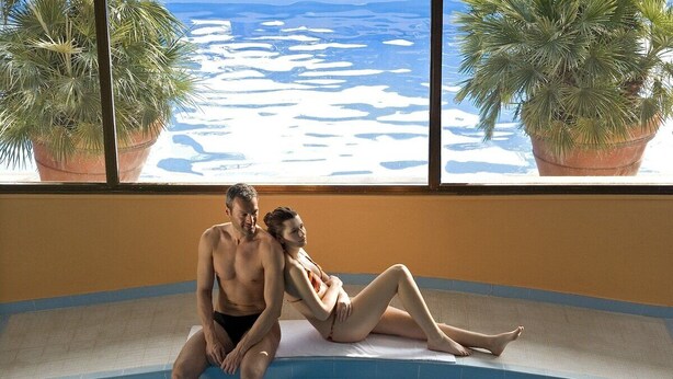 Una pareja sentada al borde de una piscina interior con vistas a un paisaje nevado a través de amplias ventanas.
